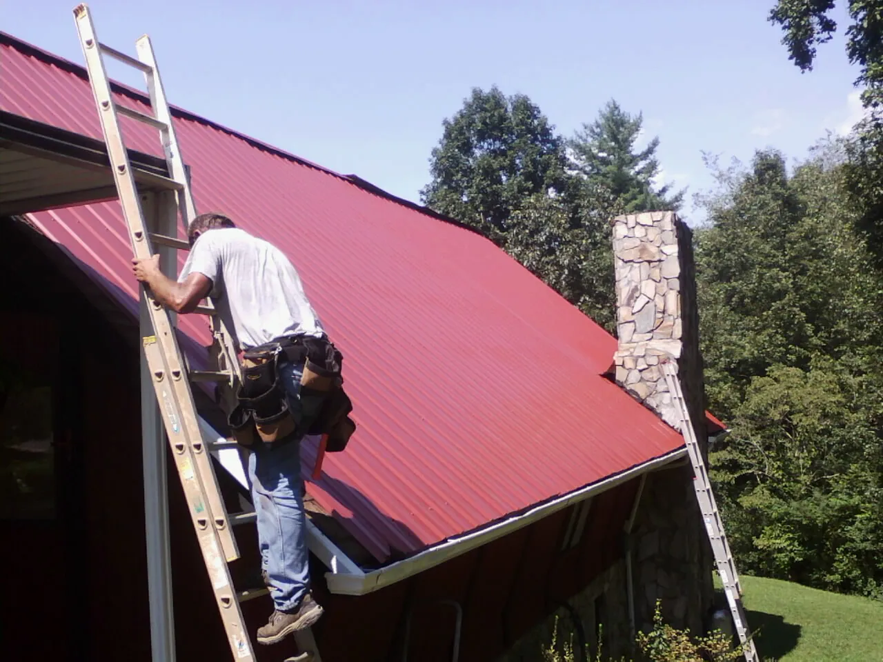 Roofer on a ladder beside a red metal roof and stone chimney — Soffit & Fascia Repair by Roofing Brothers in Logansport
